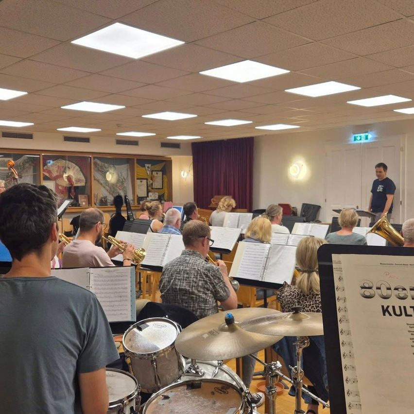 Die Probe einer Brassband in einem Saal mit Noten und Instrumenten. Ein Mann steht vorne und leitet die Gruppe.