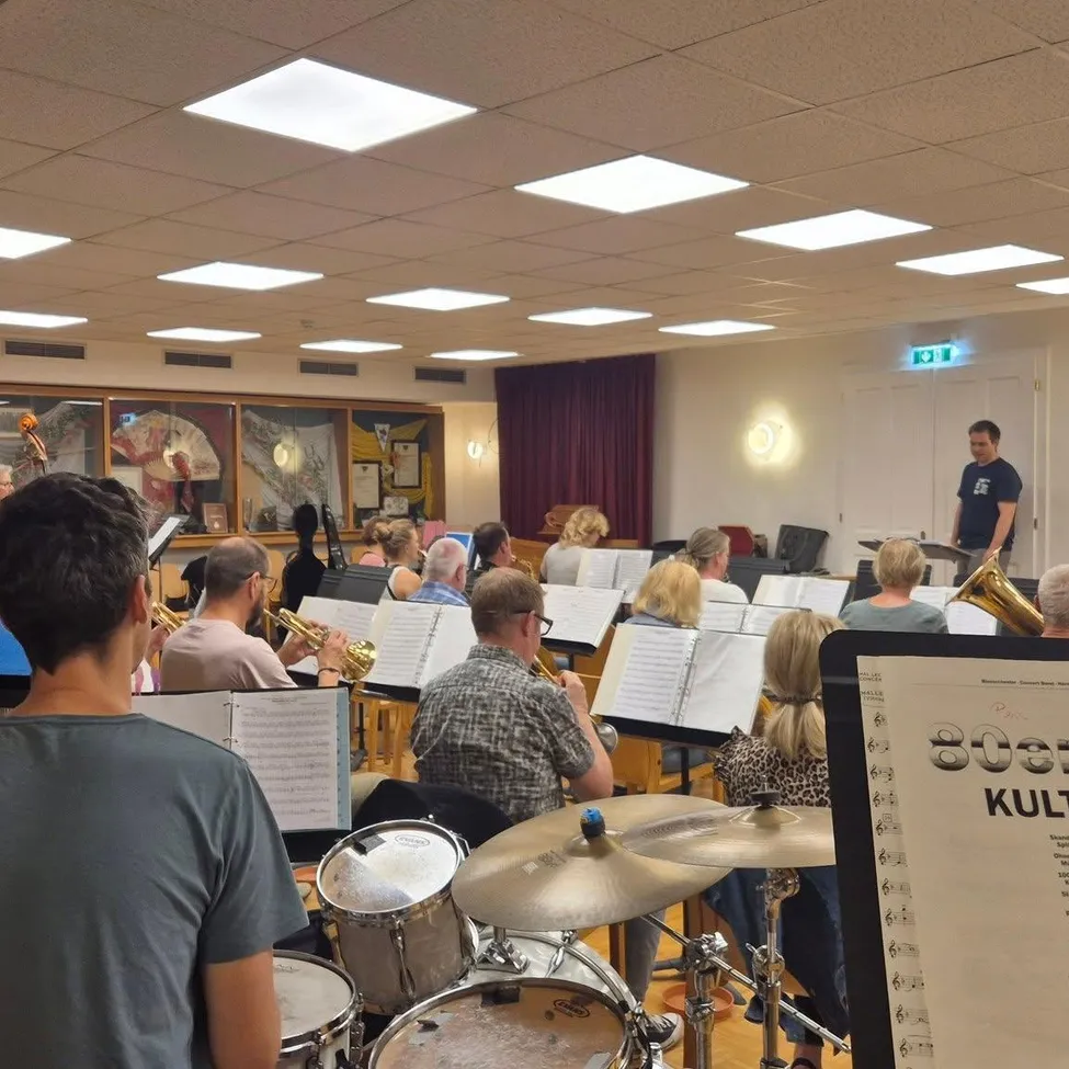 Die Probe einer Brassband in einem Saal mit Noten und Instrumenten. Ein Mann steht vorne und leitet die Gruppe.