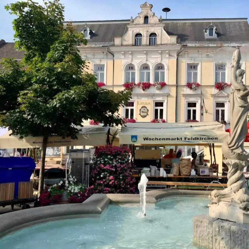 Ein Springbrunnen mit einer Statue vor einem Gebäude mit vielen Fenstern und Blumenkästen. Es gibt Marktstände mit Vordächern aufgebaut.