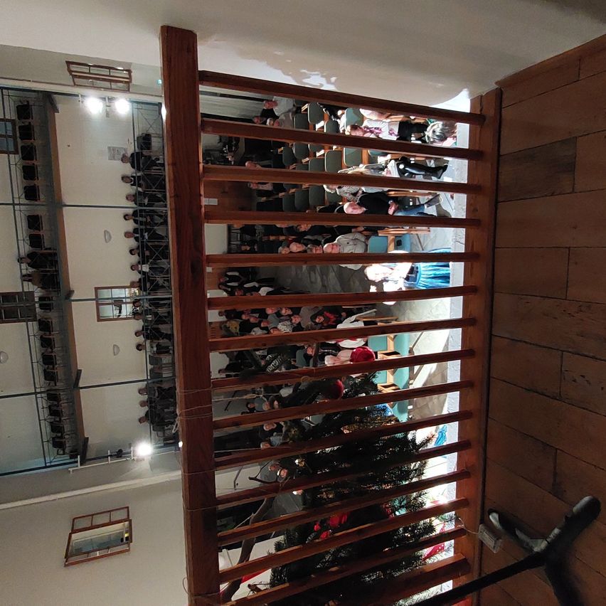 Von einem Balkon aus bietet ein hölzerner Handlauf Blick auf einen Raum mit einem Weihnachtsbaum. Unten sitzen Menschen auf Stühlen. Der Raum hat weiße Wände, Holzböden und Glasfenster.