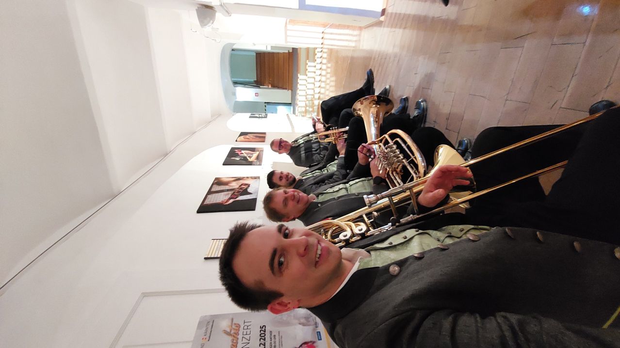 Vier Musiker, die Blechblasinstrumente spielen, lächeln für ein Foto in einem Korridor. Hinter ihnen hängt ein Plakat für ein Konzert an der Wand.