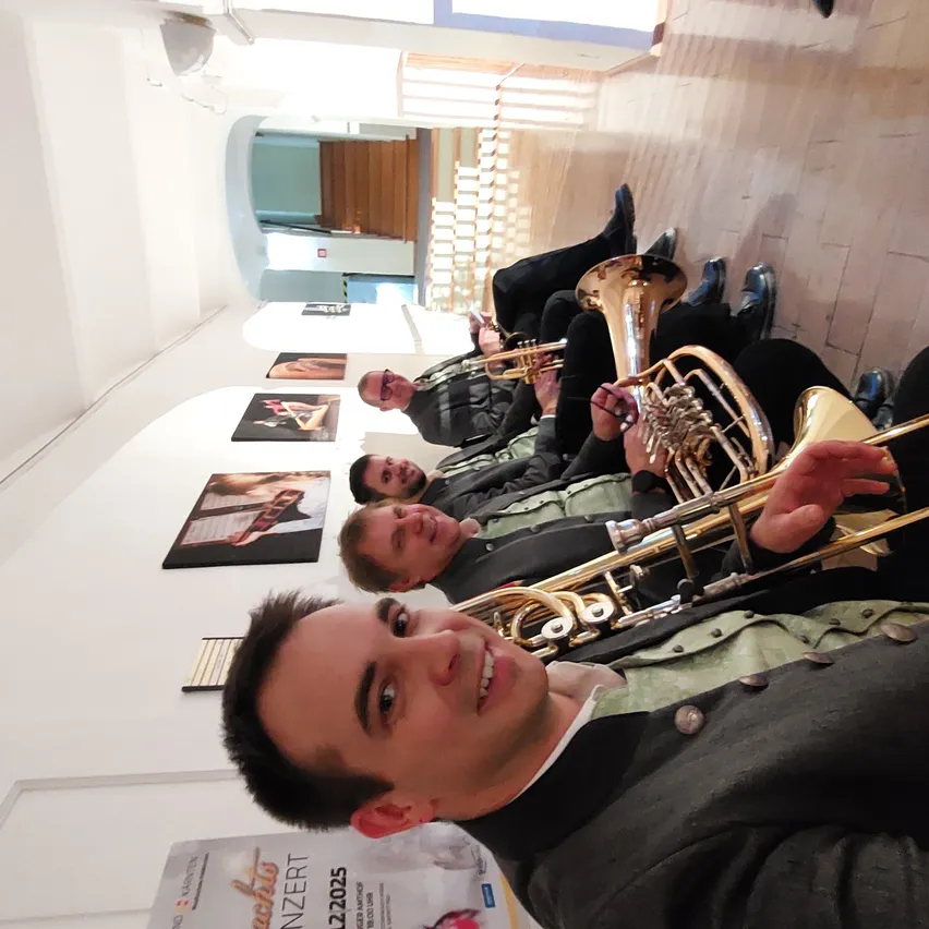 Vier Musiker, die Blechblasinstrumente spielen, lächeln für ein Foto in einem Korridor. Hinter ihnen hängt ein Plakat für ein Konzert an der Wand.