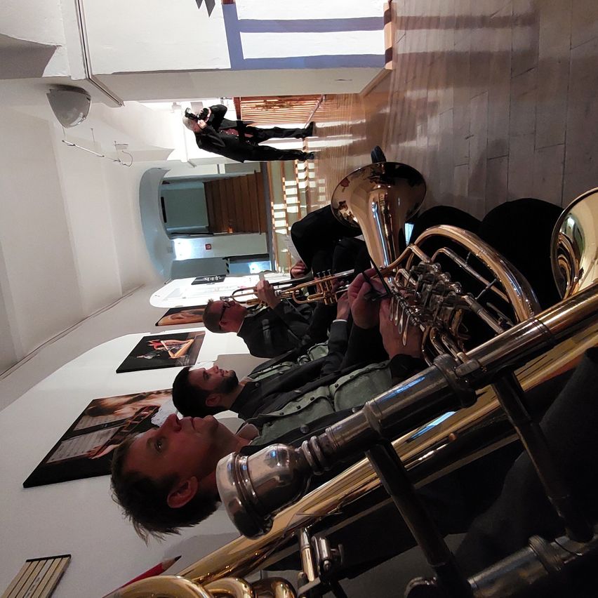 Vier Männer in einem Musikraum spielen Blechblasinstrumente. Sie stehen vor der Kamera. Ein Mann im Hintergrund macht ein Foto von den Musikern.