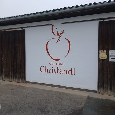 Ein weißes Gebäude mit zwei Holztüren hat ein großes rotes Apfellogo und die Worte 'OBSTBAU Christandl' darauf gemalt.