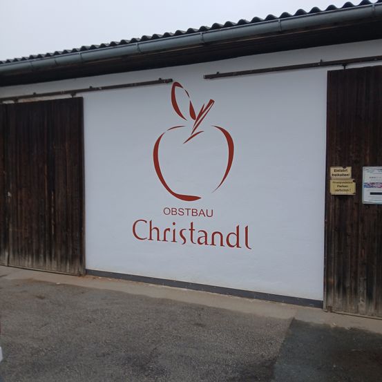Ein weißes Gebäude mit zwei Holztüren hat ein großes rotes Apfellogo und die Worte 'OBSTBAU Christandl' darauf gemalt.