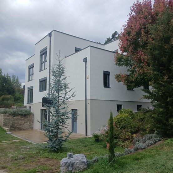 Ein modernes, weißes Haus mit mehreren Fenstern und einem kleinen Garten davor. Das Haus ist von viel Grün umgeben, darunter Bäume und Büsche. Ein Felsbrocken befindet sich im Grasbereich.