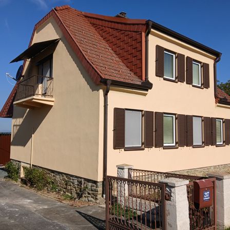 Ein Haus mit rotem Dach und beigefarbenen Wänden, mehrere Fenster mit braunen Fensterläden und einem kleinen Balkon. Ein Briefkasten befindet sich in der Nähe des Tors.