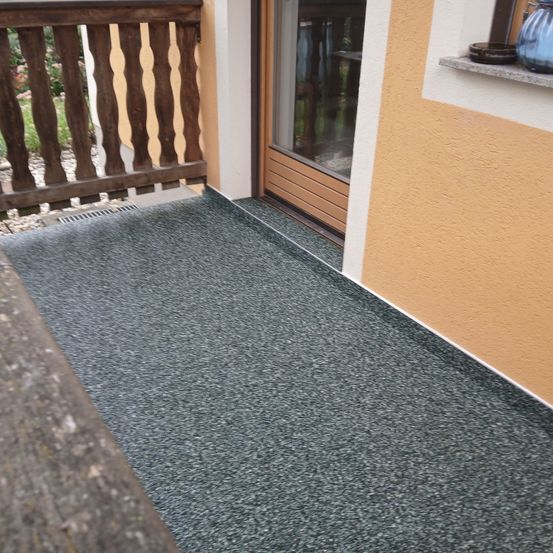 Ein Balkon mit hölzerne Balustrade und grauem Teppichboden führt zu einer Glastür mit hölzerne Rahmen. Die Wand ist gelb gestrichen.