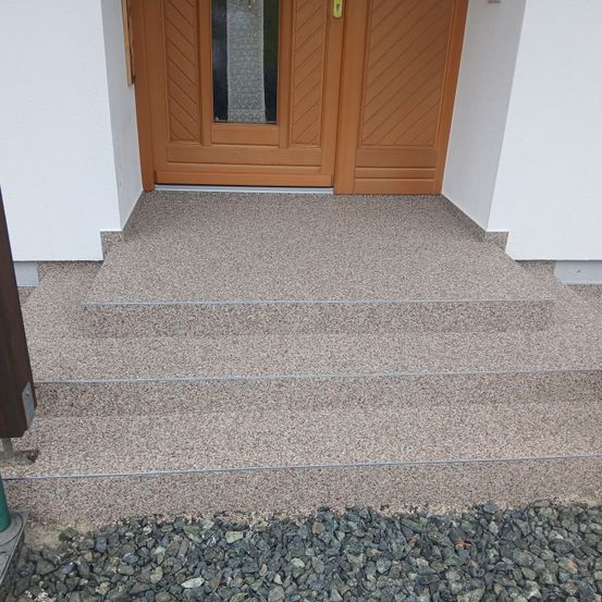 Eine braune Holztür mit Glasscheiben führt zu einem gepflasterten Weg mit drei Stufen und einem Kiesrand.