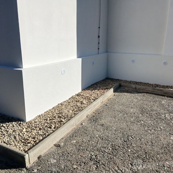 Eine weiße Gebäudeecke mit Kiesbegrenzung auf einer Seite und einem Betonrand auf der anderen. Kleine runde Elemente sind in die Wände eingebettet.