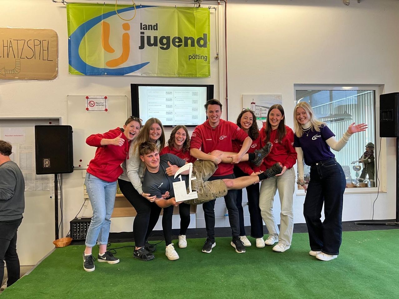 Eine Gruppe junger Menschen in roten und grauen Shirts lächelt und posiert für ein Foto. Eine Person hält ein großes Schild mit der Nummer 1. Sie stehen auf grünem Teppich. Ein Banner mit dem Text land jugend potting hängt an der Wand.