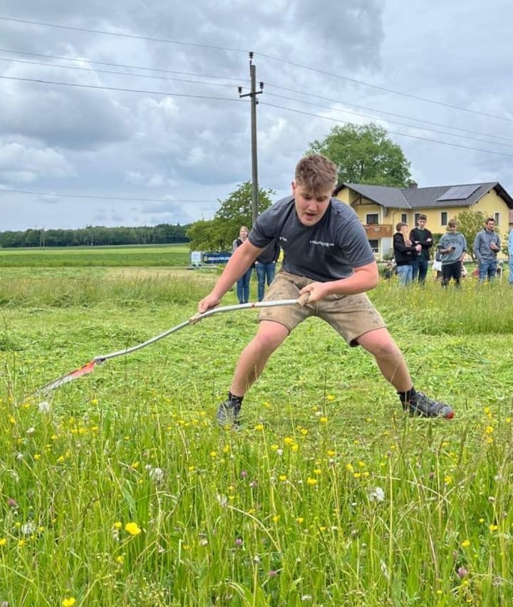 Ein Mann mäht ein grasiges Feld unter einem bewölkten Himmel, während mehrere Personen im Hintergrund zusehen.