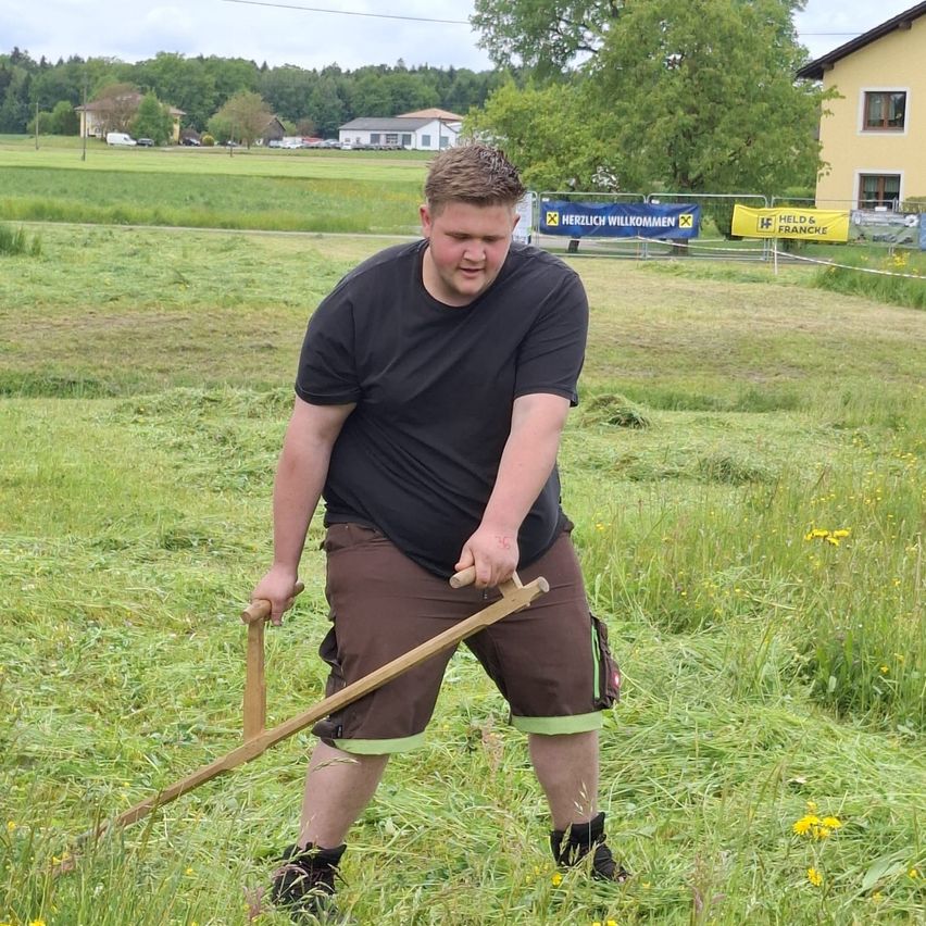 Ein Mann in einem schwarzen T-Shirt und braunen Shorts hält einen Holzstab und schneidet Gras in einem Feld mit gelben Blumen und Bäumen.