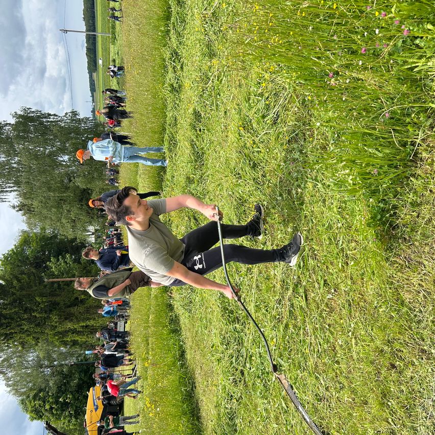 Eine Gruppe von Menschen in einem Grasfeld wartet auf ihren Einsatz bei einer Veranstaltung. Ein Mann beugt sich vor und hält eine lange Seil.