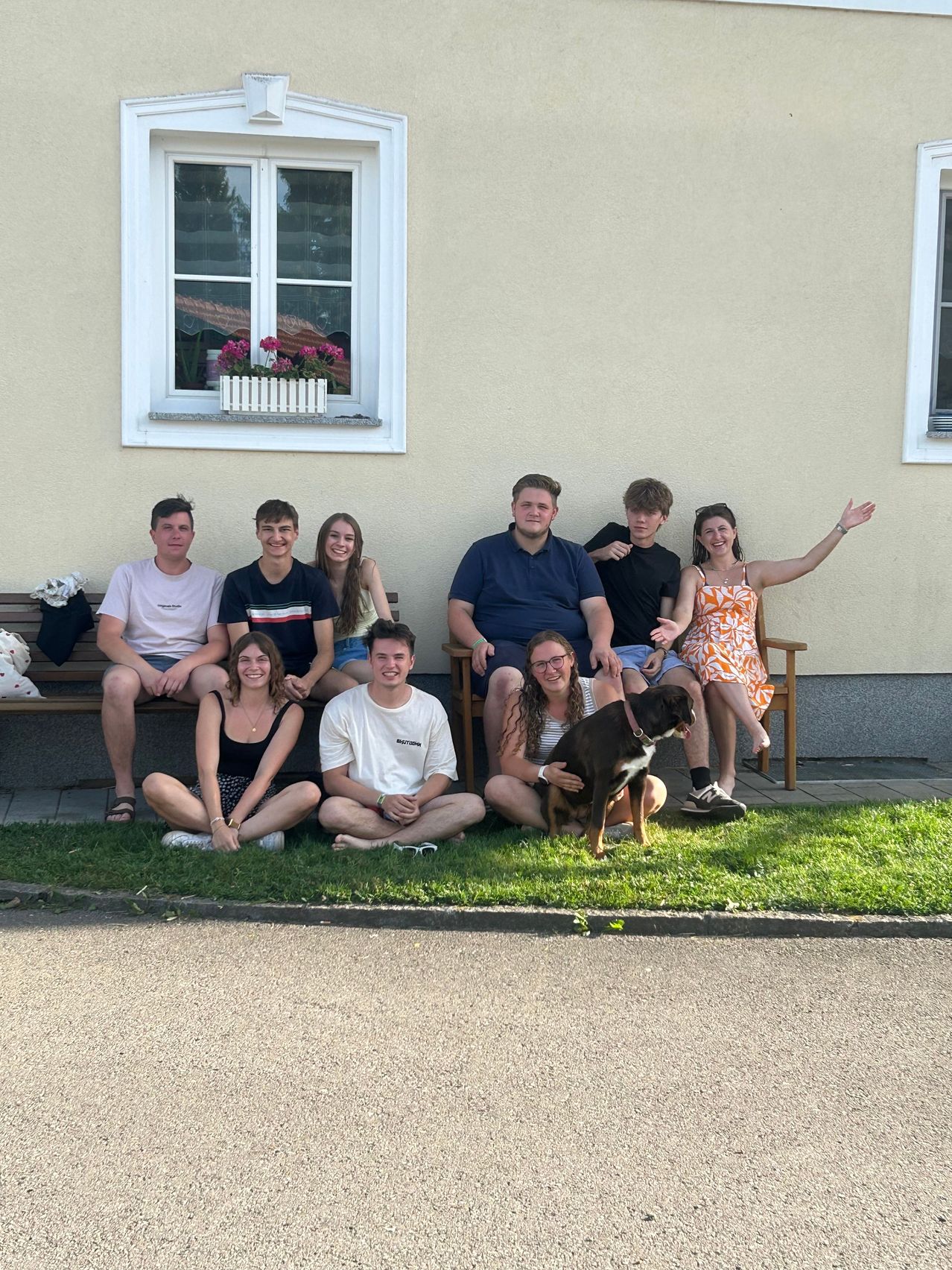 Eine Gruppe von Menschen, darunter Männer und Frauen, sitzt auf einer Bank vor einem Haus mit einem Hund vor ihnen. Sie lächeln und posieren für ein Foto.