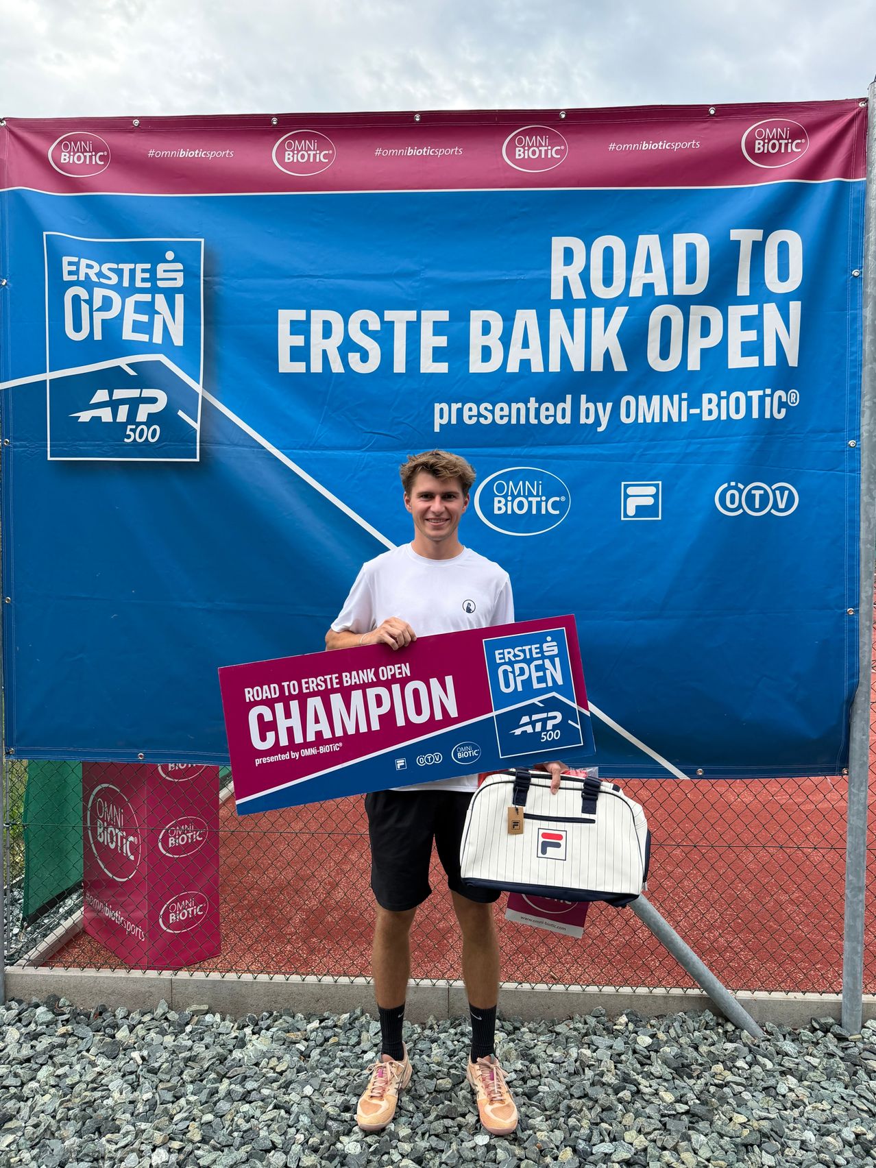 A young man stands in front of a blue banner with the text 'Road to Erste Bank Open' and holds a sign that reads 'Champion'. He is smiling and holding a tennis bag.