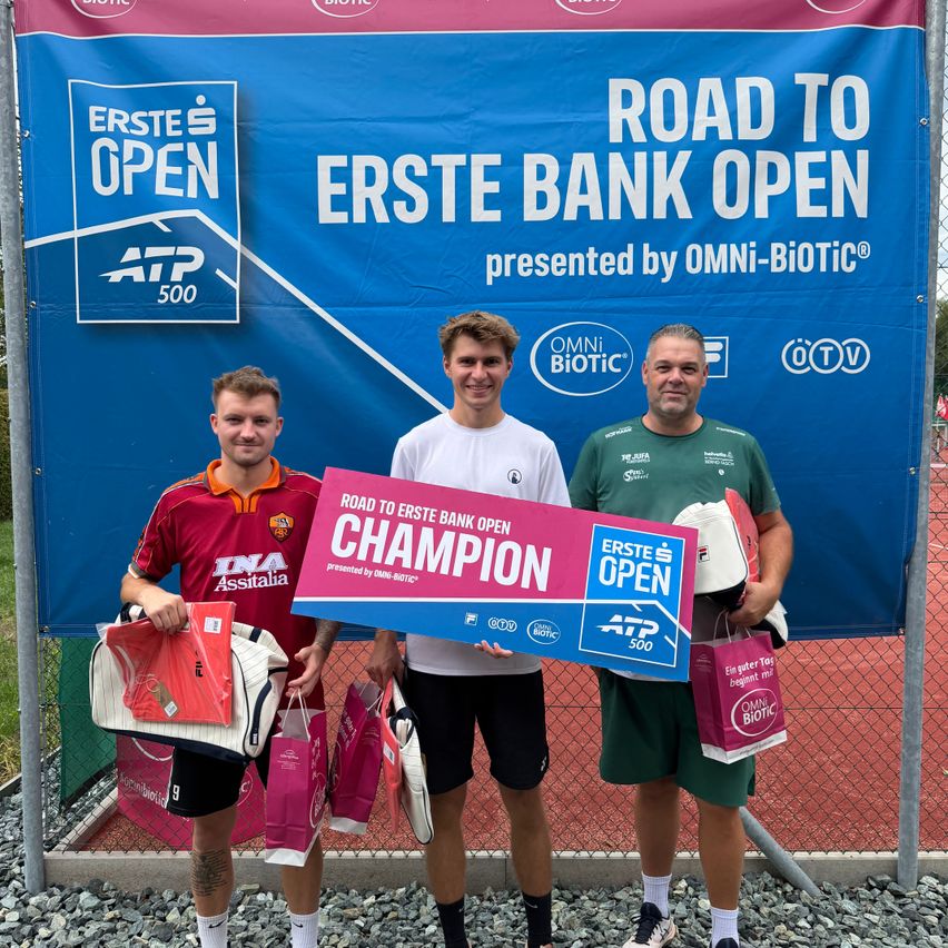 Three men stand in front of a banner for the Erste Bank Open, holding a sign that says 'Road to Erste Bank Open Champion'. The man on the left holds a bag, the middle man holds the sign, and the right man holds a bag and a racket.