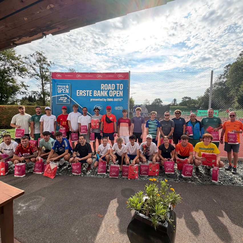 A group of tennis players pose for a photo with gift bags in front of a banner that reads 'Road to Erste Bank Open.' They are standing on a gravel surface with a fence and trees in the background.