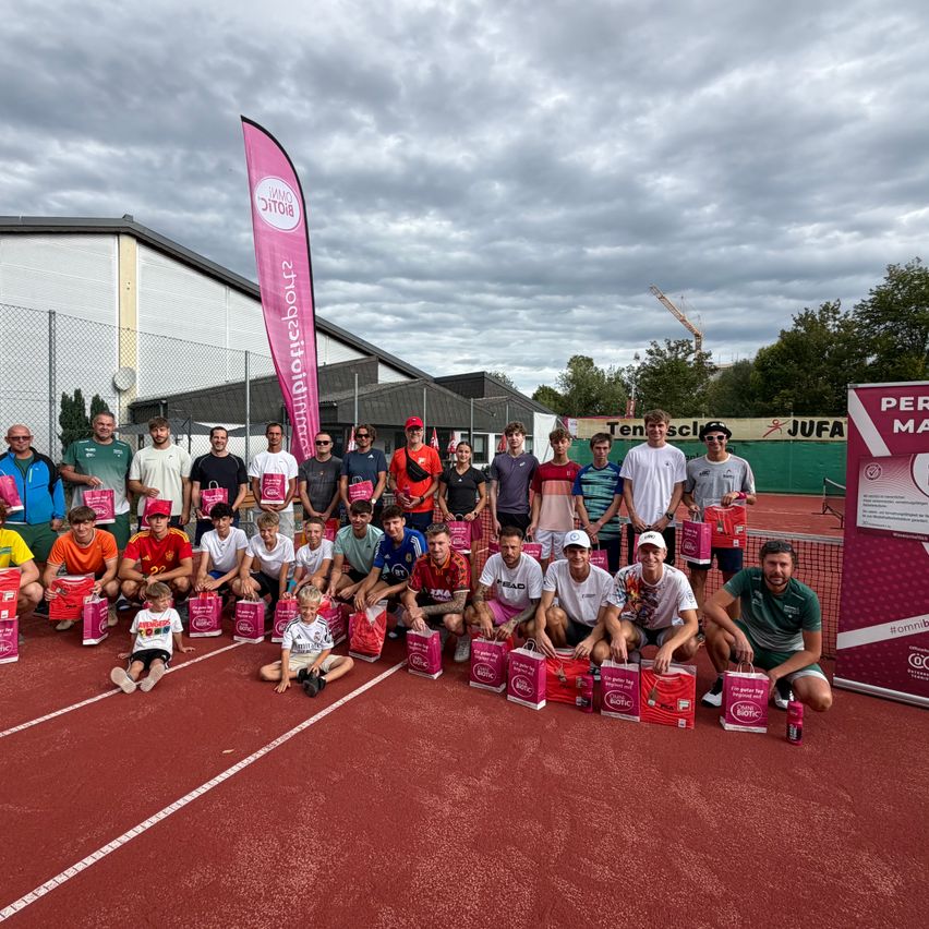 A group of people, including adults and children, are posing for a photo on a red track with a cloudy sky in the background. They are holding pink bags.