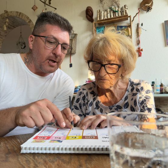 Ein Mann und eine ältere Frau sitzen an einem Tisch und arbeiten an einem Notizbuch mit farbigen Aufklebern. Der Mann konzentriert sich auf das Notizbuch, während die Frau zusieht. Hinter ihnen befindet sich eine Wand mit verschiedenen dekorativen Gegenständen.