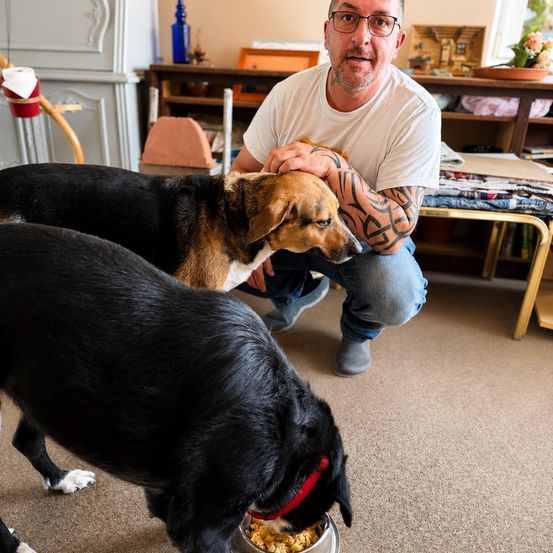 Ein Mann mit Tätowierungen kniet auf dem Boden und streichelt einen schwarz-braunen Hund, während ein anderer schwarzer Hund aus einem Napf frisst.