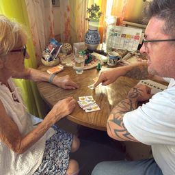 Eine ältere Frau und ein Mann mit Tattoos sitzen an einem Tisch und spielen ein Kartenspiel. Der Tisch ist mit verschiedenen Gegenständen übersät.
