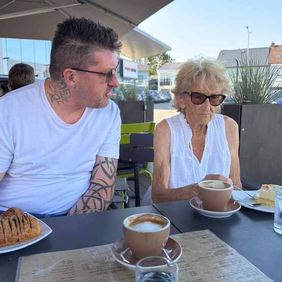 Ein Mann und eine ältere Frau sitzen an einem Tisch vor einem Café und genießen Kaffee und Gebäck. Der Mann hat Tattoos und die Frau trägt eine Sonnenbrille.
