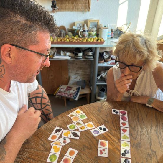 Ein Mann und eine Frau sitzen an einem Tisch und spielen ein Kartenspiel mit Obstbildern. Der Mann schaut die Frau an, während sie etwas isst.