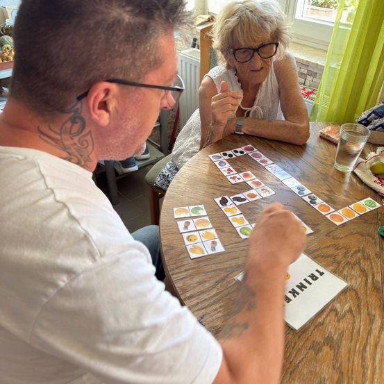 Ein Mann und eine ältere Frau sitzen an einem Tisch und spielen Scrabble. Der Mann hält einen Stift und zeigt auf das Brett, während die Frau lächelt und einen Daumen hochhält.