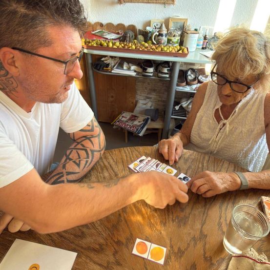 Ein Mann und eine Frau sitzen an einem Tisch und spielen ein Kartenspiel. Der Mann hat Tattoos an den Armen und die Frau trägt eine Brille. Auf dem Tisch liegen Karten, ein Glas Wasser und ein Buch. Hinter ihnen steht ein Regal mit verschiedenen Gegenständen, darunter eine Schale mit Birnen.
