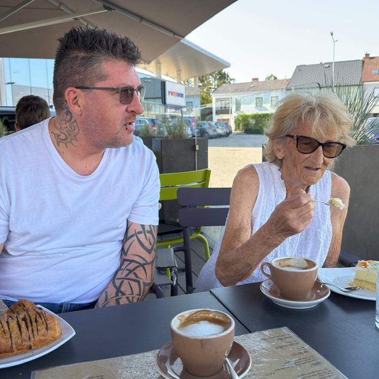 Ein Mann mit Tätowierungen und eine Frau mit Brille sitzen an einem Tisch in einem Café und essen und trinken.