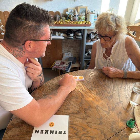 Ein Mann und eine ältere Frau sitzen an einem Holztisch und spielen ein Kartenspiel. Der Mann hat ein Tattoo am Hals.