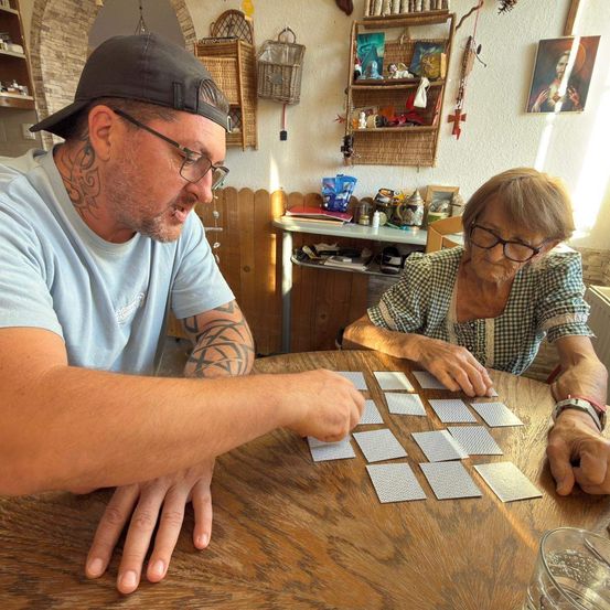 Ein Mann und eine ältere Frau sitzen an einem Holztisch und spielen ein Kartenspiel, umgeben von einem gemütlichen Raum mit verschiedenen Gegenständen an der Wand.
