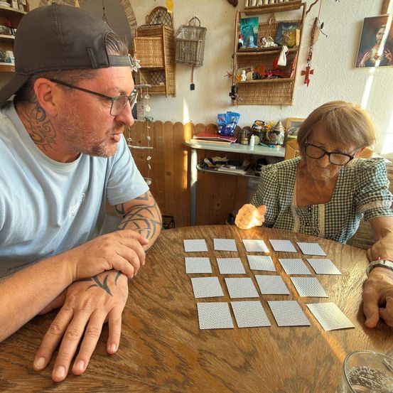 Ein Mann und eine ältere Frau sitzen an einem Tisch und spielen ein Kartenspiel. Der Mann hat Tattoos an den Armen, und die Frau trägt eine Brille. Der Tisch ist mit Karten bedeckt, und der Raum hat eine rustikale Einrichtung.