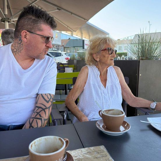 Ein Mann und eine ältere Frau sitzen an einem Tisch in einem Café im Freien, beide mit Sonnenbrillen. Der Mann hat Tattoos am Hals und am Arm. Die Frau hat eine Tasse und Untertasse vor sich.