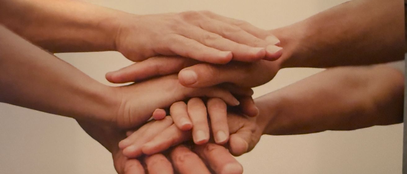 Mehrere Hände sind ineinander verschlungen und symbolisieren Einheit und Zusammenarbeit. Das Bild zeigt eine Nahaufnahme von Fingern verschiedener Personen.