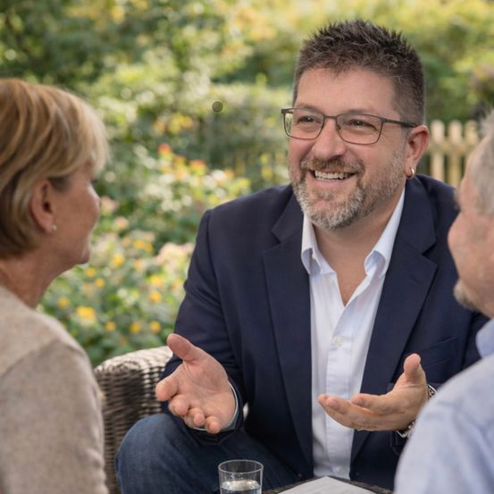 Ein Mann in Anzug und Brille lächelt und gestikuliert, während er mit zwei Personen in einem Garten spricht.