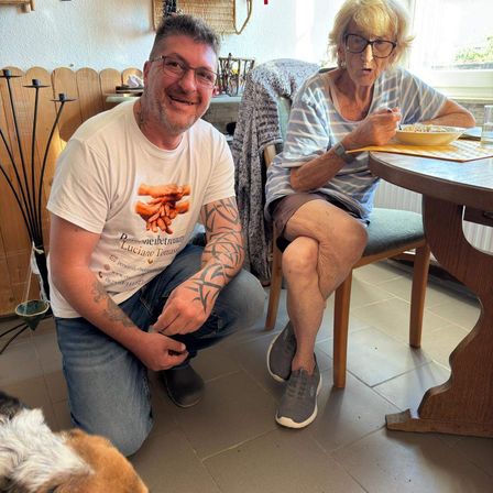 Ein Mann mit Tätowierungen kniet neben einer älteren Frau, die am Tisch sitzt und beide lächeln. Die Frau isst aus einer Schüssel, und ein Hund ist in der Nähe.