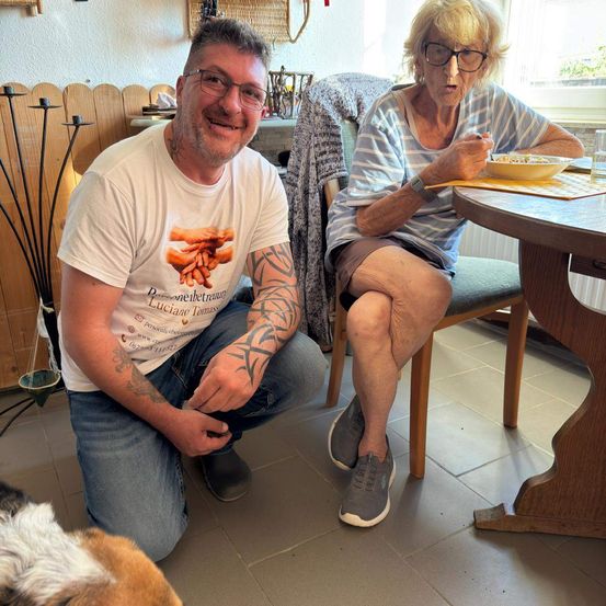 Ein Mann mit Tätowierungen kniet neben einer älteren Frau, die am Tisch sitzt und beide lächeln. Die Frau isst aus einer Schüssel, und ein Hund ist in der Nähe.