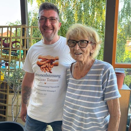 Ein Mann und eine ältere Frau stehen lächelnd eng beieinander in einem Garten. Der Mann trägt ein weißes T-Shirt mit einer grafischen Gestaltung, und die Frau trägt ein blau-weiß gestreiftes Shirt. Hinter ihnen steht eine Holzstruktur und eine Topfpflanze.
