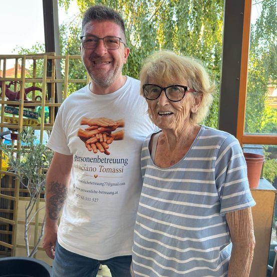 Ein Mann und eine ältere Frau stehen lächelnd eng beieinander in einem Garten. Der Mann trägt ein weißes T-Shirt mit einer grafischen Gestaltung, und die Frau trägt ein blau-weiß gestreiftes Shirt. Hinter ihnen steht eine Holzstruktur und eine Topfpflanze.