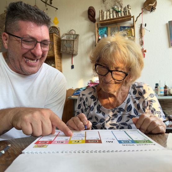 Ein Mann und eine ältere Frau sitzen an einem Tisch, lächeln und schauen auf eine farbenfrohe Tabelle. Der Mann zeigt mit einem Stift auf die Tabelle.