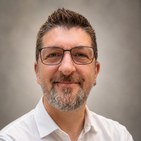Ein Mann mit Bart und Schnurrbart, der eine Brille und ein weißes Hemd trägt, lächelt und schaut nach vorne.