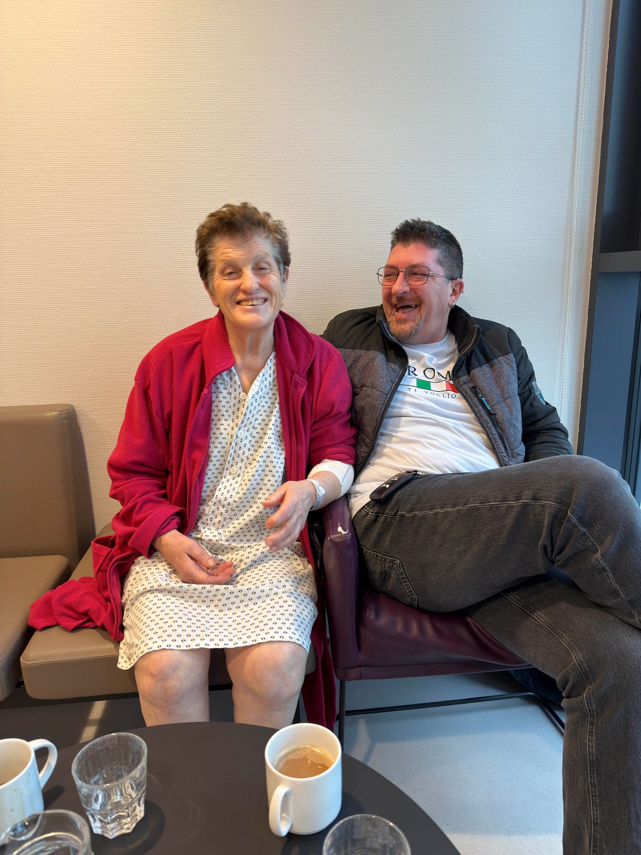 Eine lächelnde ältere Frau in einem Krankenhauskleid und ein Mann in Freizeitkleidung sitzen zusammen auf einem Stuhl in einem Krankenhauszimmer.