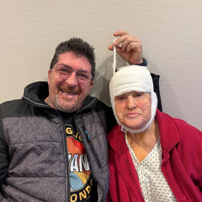 Ein lächelnder Mann mit Brille und Jacke hält den Kopf einer Frau mit einem Verband daran. Die Frau trägt einen roten Mantel und einen weißen Verband auf dem Kopf.