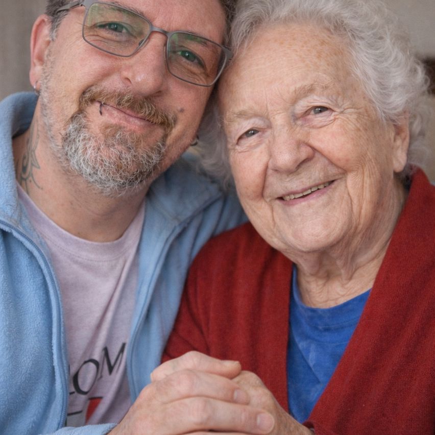 Eine ältere Frau mit weißen Haaren lächelt und hält die Hand eines Mannes, der eine Brille trägt.