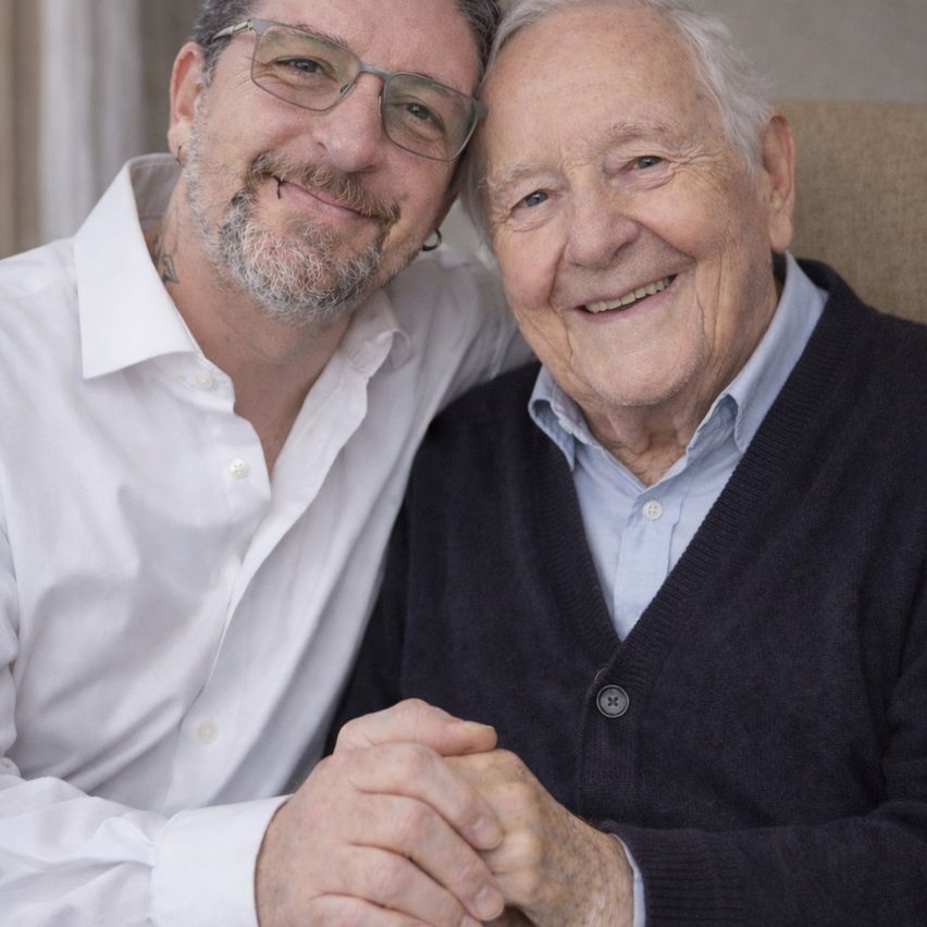 Zwei Männer, einer älter mit weißen Haaren und einem schwarzen Cardigan, lächeln und halten Händchen mit einem jüngeren Mann in einem weißen Hemd.