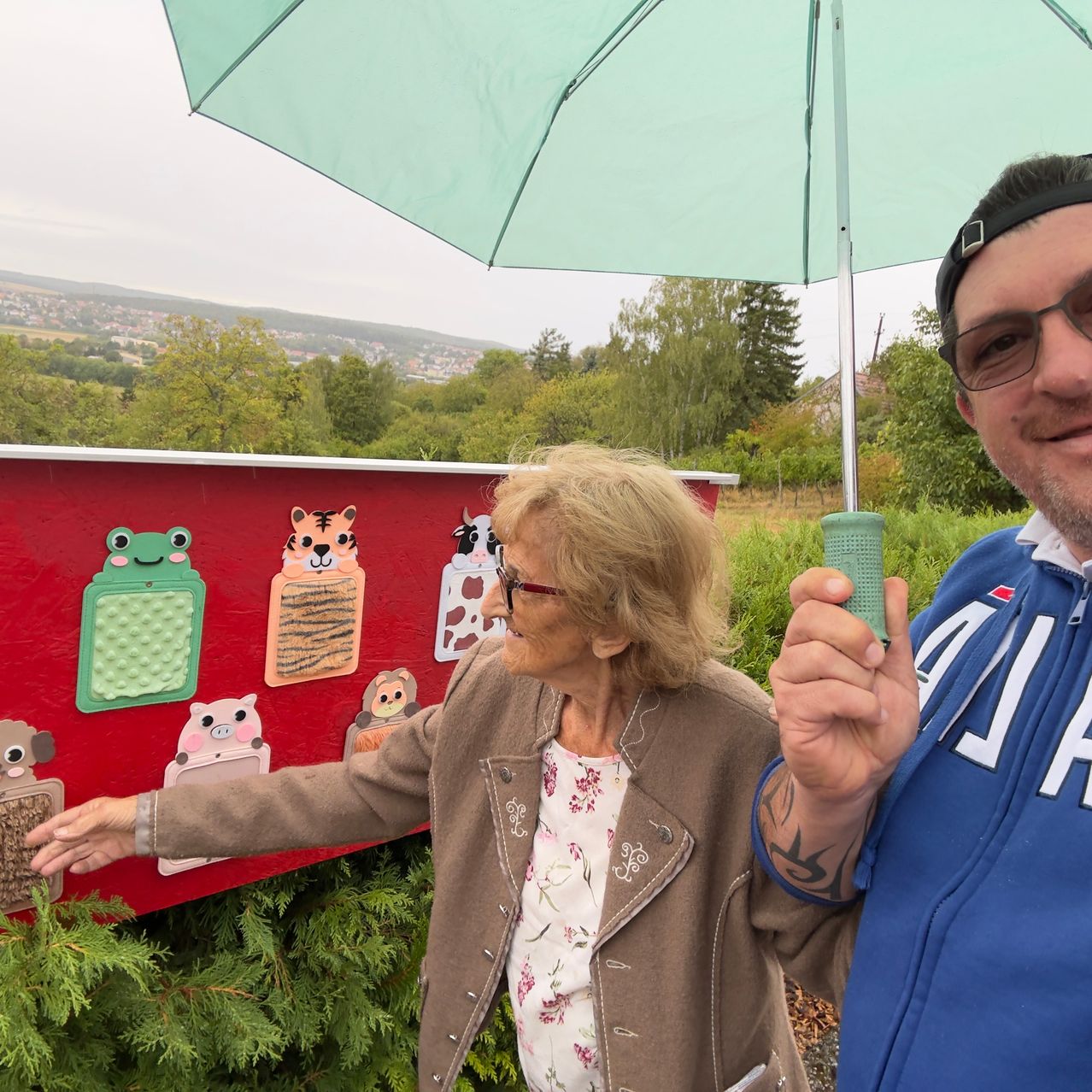 An older woman and a man stand under an umbrella near a red wall with animal illustrations. The woman touches the wall while the man holds a green object.