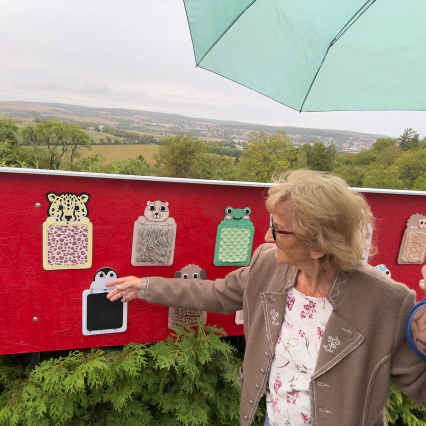 An older woman with glasses stands under an umbrella, pointing at a red wall with tactile animal illustrations.