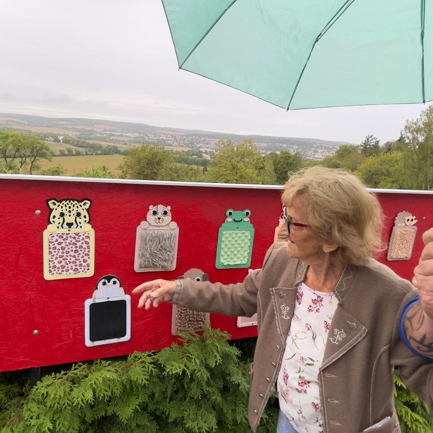 An elderly woman stands under a green umbrella, pointing at a red board with various tactile animal images.